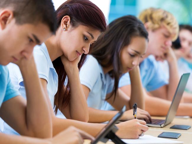 Row of private high school students work on assignment in class. They are writing or using laptops or digital tablets. They are concentrating as they study. They are wearing school uniforms.