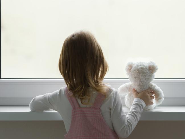 Little girl holding white teddy bear and standing alone at window and looking out from home. Back view. Waiting concept. Close up.