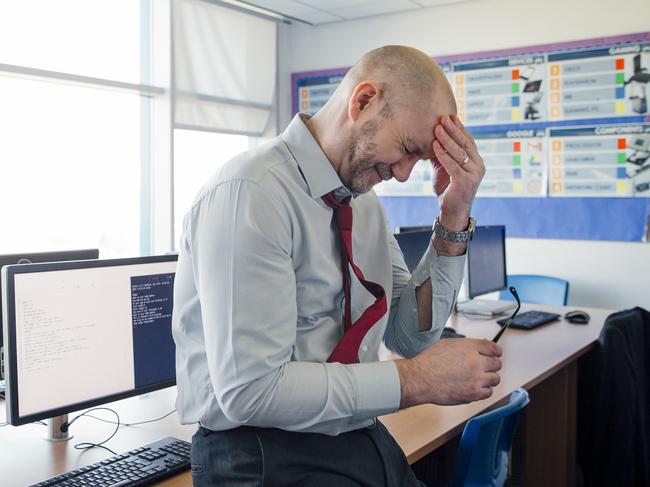 A teacher sits on the edge of a desk in an empty classroom, he is stressed