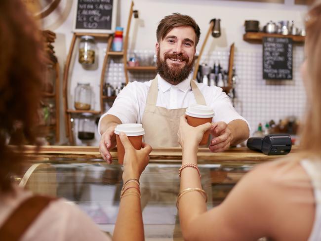 Friendly young man with beard standing behind the cafe counter and serving coffee for two women. Young man working in restaurant, helping customer with coffee.