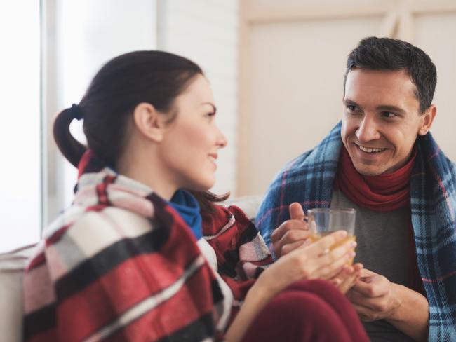 The couple is sitting on the couch wrapped in blankets. Man and woman are sick. A man gives a woman hot medicinal tea in a glass cup. Woman smiling RendezView