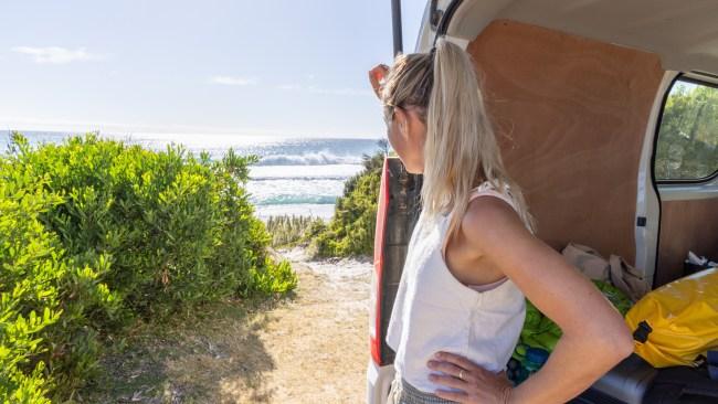 A woman with a ponytail is standing by the open trunk of a van, enjoying the view of the ocean and beach. The van is filled with various belongings, and the scenic coastline is visible in the background.