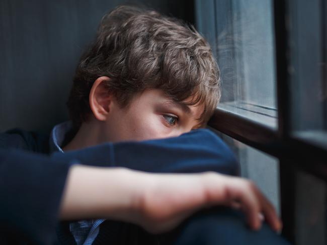 Pensive sad boy teenager with blue eyes in a blue shirt and jeans sitting at the window and closes his face with his hands.