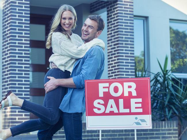 Couple in front of a new home. They are standing next to a for sale sign. They are both wearing casual clothes and embracing. They are smiling and he has a beard. He is lifting his wife off the ground with joy. The house is contemporary with a brick facade. The front door is also visible. Copy space