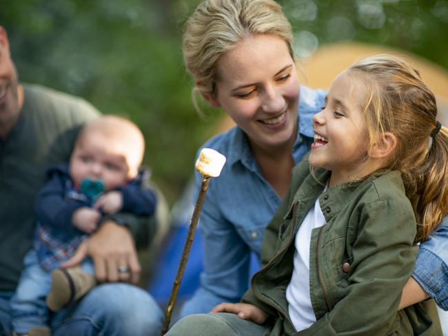 A family roasts marshmallows while on a camping adventure. The mother is holding her daughter while the father holds their baby.Photo - iStockEscape 3 april 2022Kidspot family camping