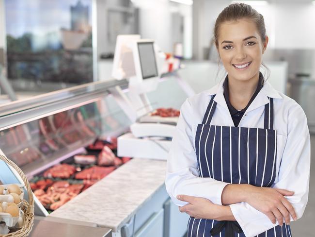 a young female butcher tends the meat counter in a butcher's shop. She is wearing a white coat and striped apron and is smiling to camera. Behind her the meat counter can be seen defocussed . butcher shop generic Savvy Shopper