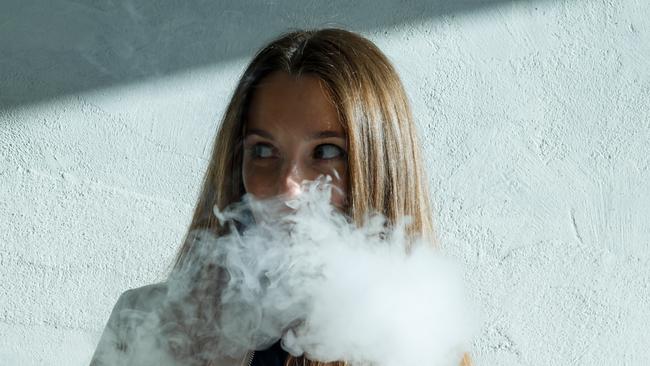 Vape teenager. Young cute girl in  casual clothes smokes an electronic cigarette near the wall outdoors in summer day. Bad habit that is harmful to health. Vaping activity.