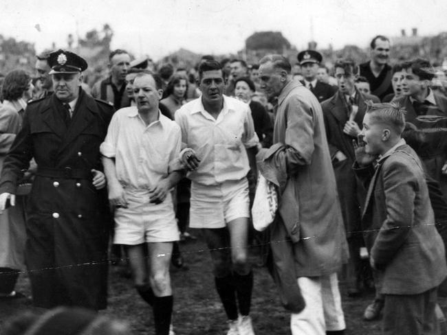 1957: Umpire Woolfe is escorted by police from the ground at an Essendon v Hawthorn VFL match. Woolfe was pelted with rubbish and hooted on one of the noisiest demonstrations ever seen at Windy Hill.