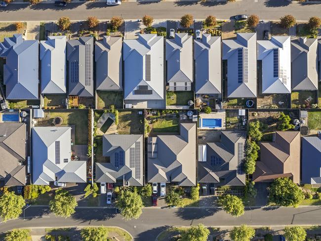 Aerial view directly above new outer suburban/semi-rural housing development with single-level housing between two streets with t-intersections and orange coloured street trees.  Mostly gray metal roofing, landscaped front and back yards, some solar panels, cars and motor vehicles parked in driveways and street, some street and parkland trees, water tanks in backyards, sheds and garages, swimming pools.  Mount Barker, South Australia; property investment housing money generic