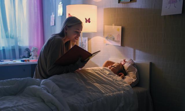 Young Loving Mother Reads Bedtime Stories to Her Little Beautiful Daughter who Goes to Sleep in Her Bed.
