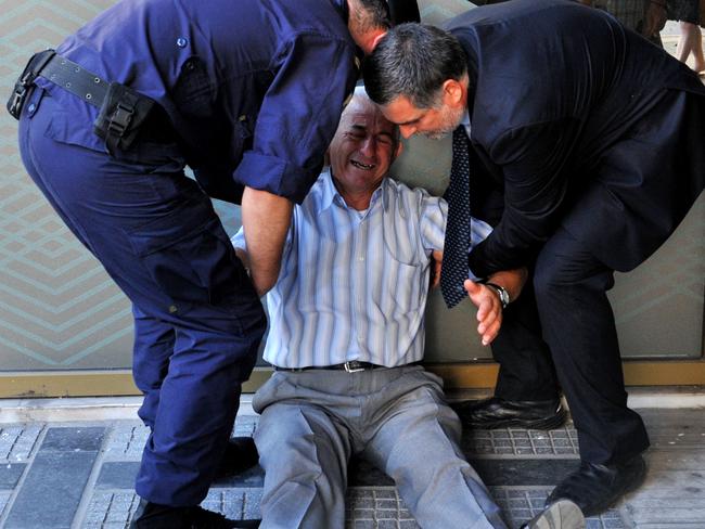 Giorgos Chatzifotiadis, assisted by an employee and a policeman, sits on the ground outside a national bank branch, as pensioners (unseen) queue to withdraw their pensions. Picture: SAKIS MITROLIDIS