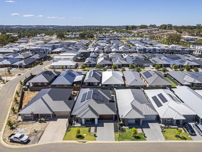 Low aerial close view new dense rural housing development, mostly grey roofing, some green landscaping, young trees. Rows of houses of various architectural styles with shades of grey roofing in rural urban sprawl, some still under construction.  Tightly-packed houses with small front and back yards and few if any trees. Some solar panels installed. Mt Barker near Adelaide, South Australia