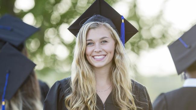 A multi-ethnic group of university students are outdoors at a graduation ceremony. They are wearing graduation robes and caps. They are walking away from the camera, and a Caucasian woman is turned to smile at the camera while holding her diploma.