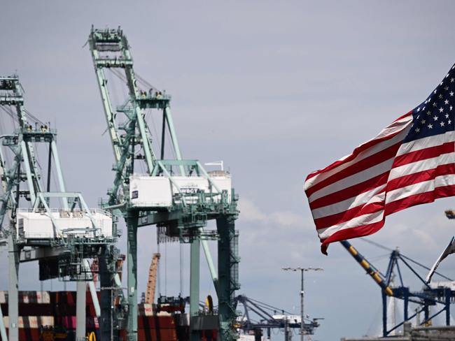 (FILES) The US flag blows in the wind as cranes stand above cargo shipping containers on ships at the Port of Los Angeles in Los Angeles, California on June 7, 2023. An increase in exports helped to reduce the overall US trade deficit in August to the smallest since late 2020, according to government data released on October 5, 2023. The trade gap shrank to $58.3 billion, down nearly 10 percent from July's revised figure of $64.7 billion, said the Commerce Department. (Photo by Patrick T. Fallon / AFP)