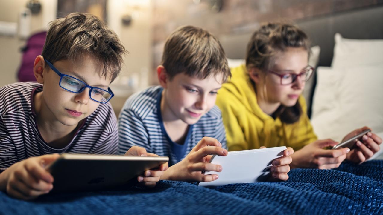 Three kids are lying on bed and playing with modern mobile devices – tablets and smartphone. Nikon D850