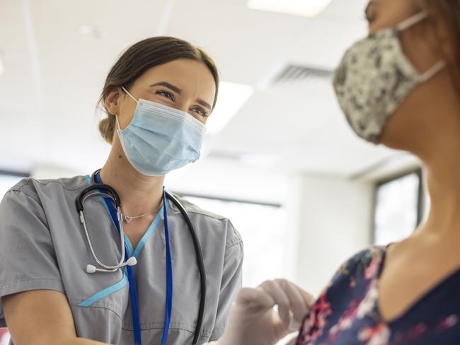 Shot focused on a kind looking nurse wearing scrubs, a stethoscope, white rubber gloves and a protective face mask. She is smiling at the nervous patient with her eyes. The nurse is prepping the patients arm before she injects her with the COVID-19 vaccine.