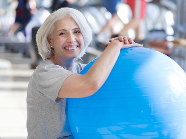 Finishing training. Pleasant beautiful senior woman smiling and keeping fit ball while sitting on a floor in a gym. Healthy baby boomers and seniors, generic retirement