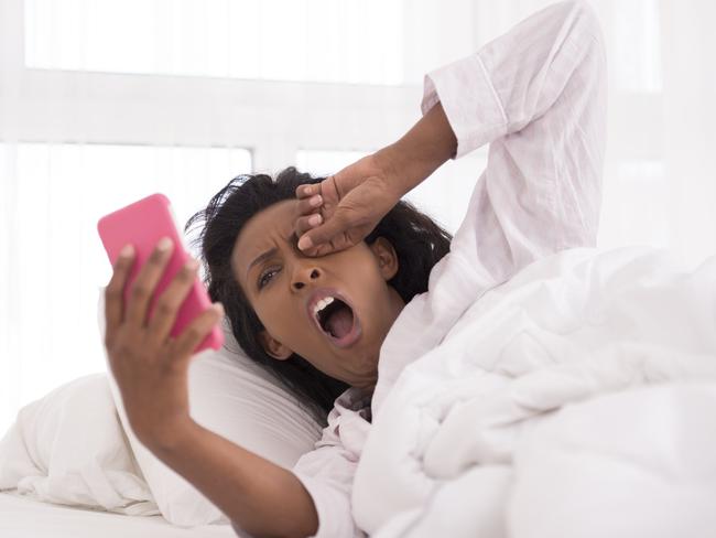 Woman yawning, waking up with smartphone in hands, texting, checking messages while rubbing her eye.