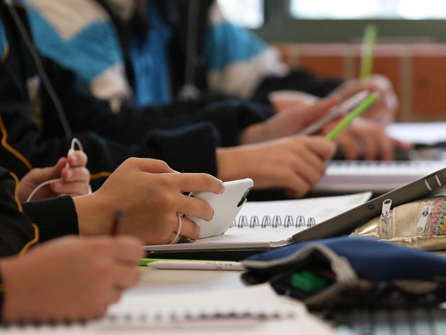 A student in uniform distracted holding using and watching a mobile phone during a lesson at high school. Books, tablets and pencil cases all visible on the desk and work space.