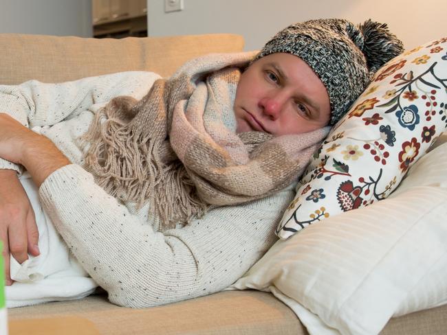 Sick man wearing scarf lying on couch at home under a blanket checking temperature. Living room with table full of medicine and pills. Man with running nose and cough. Istock