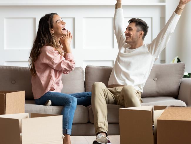 Full length happy married family couple sitting together in living room, laughing celebrating moving in new house. Overjoyed homeowners feeling excited about apartment flat mortgage purchase.