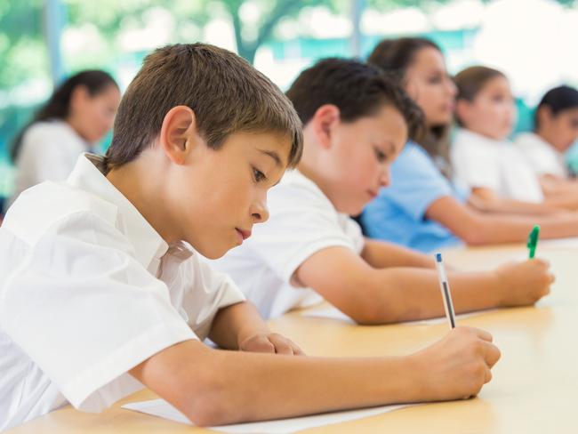 Elementary age Hispanic little boy is sitting at desk with classmates in private elementary school. He is using a pen to write on paper during exam. Classmates are also concentrating while taking test. Students are wearing white and blue school uniforms.