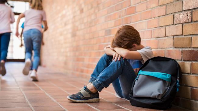 Little boy sitting alone on floor after suffering an act of bullying while children run in the background. Sad young schoolboy sitting on corridor with hands on knees and head between his legs.