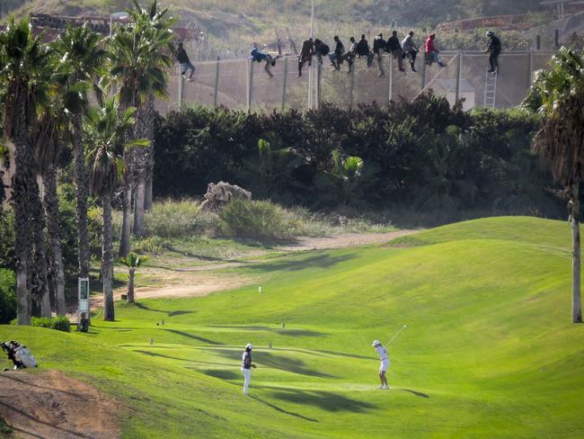 An extraordinary image shows golfers playing just metres from fence-climbing migrants. Picture: Prodein