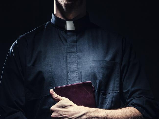 A dark high contrast image of a clergy man in a clerical collar holding a brown leather bound Bible in his arms.