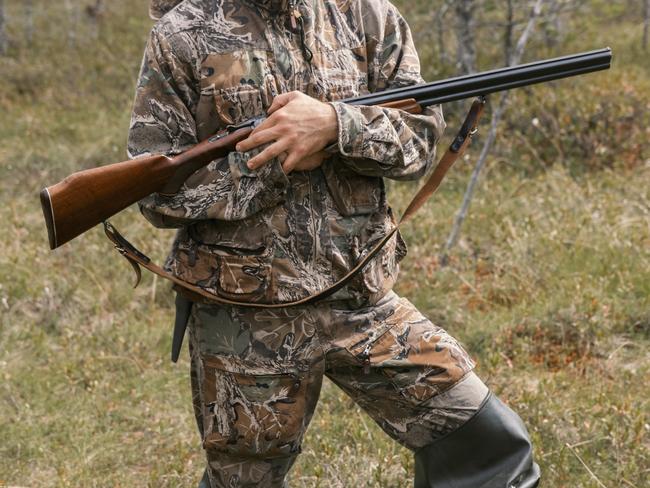 An experienced hunter in full camouflage gear steadies his rifle while navigating through a swampy area. His outfit blends seamlessly with the surrounding foliage, and the careful handling of the firearm emphasizes responsible gun control. The setting suggests a typical hunting scene, capturing a moment of anticipation and readiness in the wild.