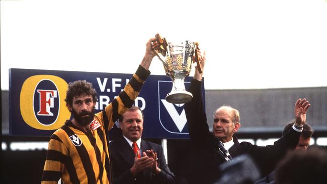 Hawthorn captain Michael Tuck and coach Allan Jeans lift the Premiership Cup (with Ted Whitten pictured in the background) after beating Geelong in the 1989 VFL Grand Final. The league became the AFL the following season.