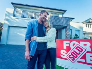 Couple standing in front of a new home. They are standing next to a for sale sign with a sold sticker. They are buying or selling this real estate. They are both wearing casual clothes and embracing. They are smiling and he has a beard. The house is new and contemporary with a brick facade. Copy space