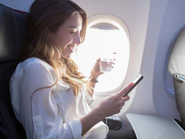 A smiling young woman sits in the window seat of her aircraft.  She holds a beverage in one hand and her smart phone in another as she enjoys the wifi.  Her earbuds rest on a tray in front of her. Picture: iStockflight mode wisdom, Amanda Woods, Escape