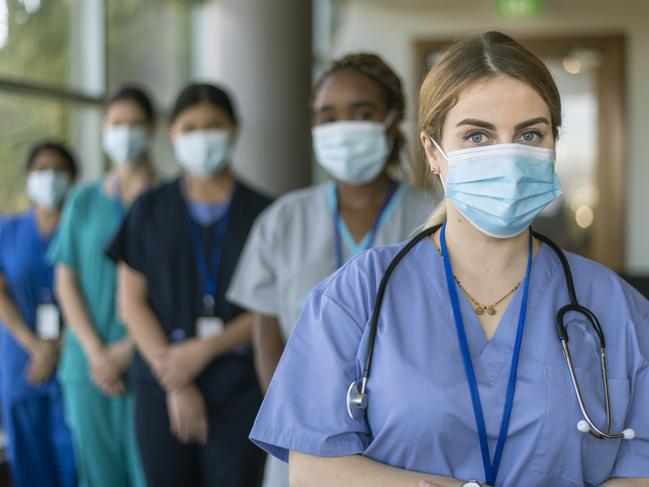 A multi-ethnic team of medical workers wearing scrubs and protective face masks look directly at the camera while posing in a line in a hospital corridor while working during the Covid-19 pandemic. Image is focused on a female doctor standing at the front of the group.