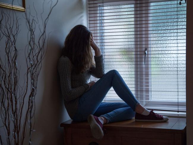 DTR914 Young female with long hair wearing jeans, hand on head in despair, sitting in darkness by a window at home. Side viewpoint.