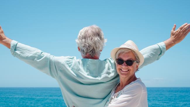 Cheerful senior woman with hat hugging her husband while looks at horizon over sea with outstretched arms. Happy retired couple enjoying summer holidays. Happy senior couple generic