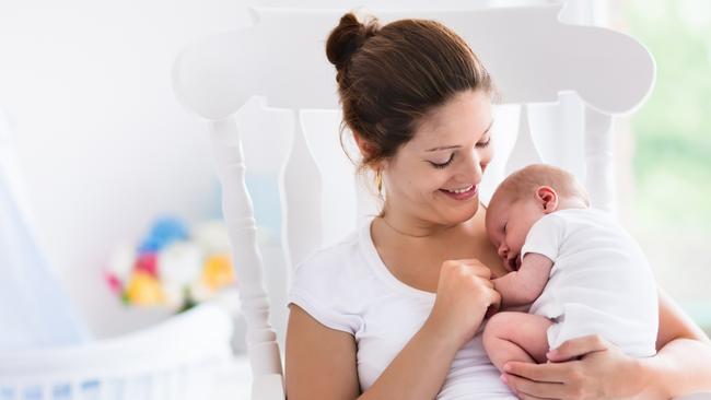 Young mother holding her newborn child. Mom nursing baby. Woman and new born boy relax in a white bedroom with rocking chair and blue crib. Nursery interior. Mother breast feeding baby. Family at home