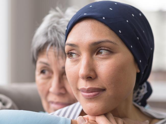 An ethnic woman wearing a headscarf and fighting cancer sits on the couch with her mother. She is in the foreground and her mom is behind her, with her arm wrapped around in an embrace, and they're both looking out the window in a quiet moment of contemplation.