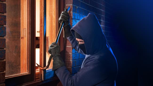 Hooded masked burglar forcing open a window in a home.