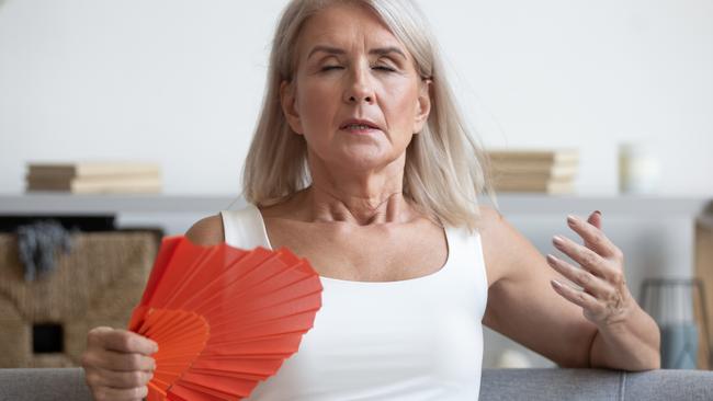 Tired mature woman suffering from heat at home, feeling uncomfortable, waving blue fan, sitting on couch, enjoying fresh air, sweaty older female cooling in hot summer weather, high temperature