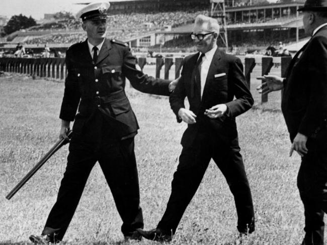 1966: Anti-whip campaigner Wally Hoysted is led away by a police officer at Flemington racecourse in Melbourne after walking onto the track with a double-barrelled shotgun. Hoysted fired a shot and threatened to use the gun on jockeys preparing for the first race of the day before being taken away. Picture: Bruce Howard/File