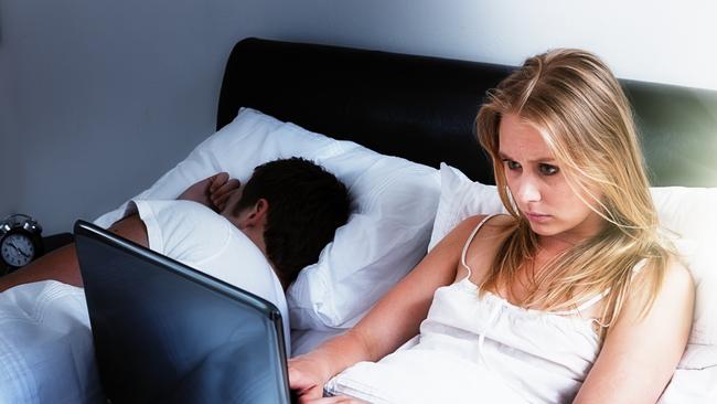 A handsome young couple lie in bed; she's intent on her laptop, either keeping up with social media, working late, or battling insomnia; he's fast asleep next to her. Istock