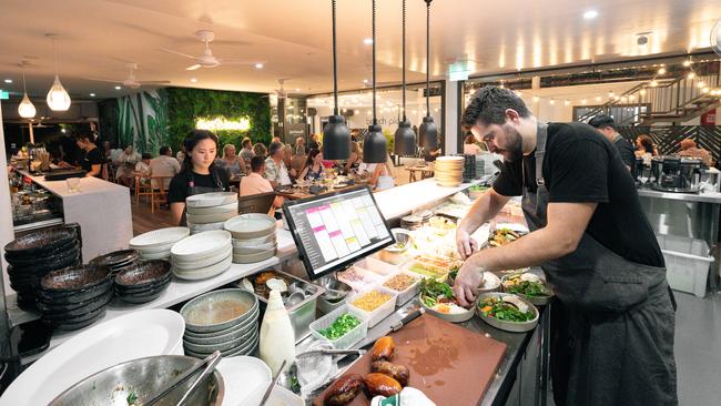 Showing the view from the kitchen, the restaurant features a large tropical leaf mural that has been painted by a local artist, and a vertical plant wall with the glowing sign 'Eastwoods'. Picture: Simone Hutchen Creative.