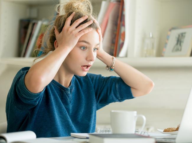 money mistakes - generic woman shocked, stressed... Portrait of an attractive woman at the table with cup and laptop, book, notebook on it, grabbing her head. Bookshelf at the background, concept photo