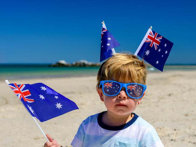 Cute smiling kid with Australian flags sitting on the sand at the beach on Australia Day