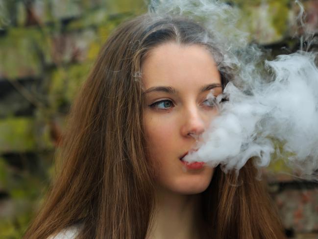 Vape teenager. Young pretty white girl in white cardigan smoking an electronic cigarette opposite destroyed brick wall on the street in the spring. Bad habit that is harmful to health.