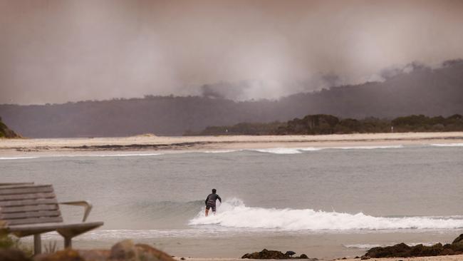 A surfer catches a wave in Mallacoota while the fires burn in the distance. Picture: David Caird