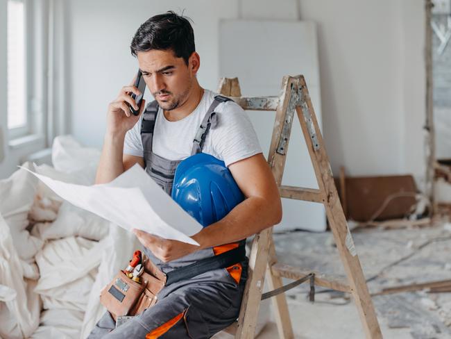 Developing Queensland - A young Caucasian male construction worker is having a phone call, while sitting down and holding the renovation plans.