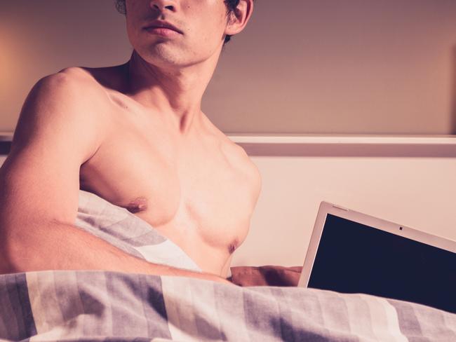 Sexy young man in in bed under a duvet is using a laptop and looking back over his shoulder