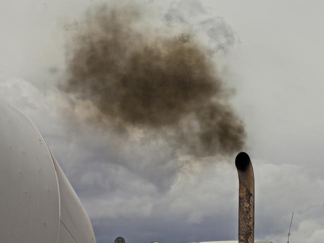 A white dump truck spews black exhaust smoke into a blue sky. Just the top of the truck is visible with the sky taking up half the image. The burning of fossil fuel by vehicles contributes to greenhouse gasses, pollution in urban city centers, and health concerns of people worldwide. Diesel fumes